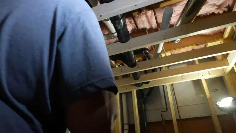 Plumber in a blue shirt looking up at supply and drain pipes running between unfinished basement joists